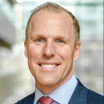 A smiling man with short blond hair and blue eyes. Wearing a dark blue plaid blazer, a light blue dress shirt, and a red patterned tie