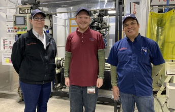 Alex stands in between two other adults in front of some factory equipment
